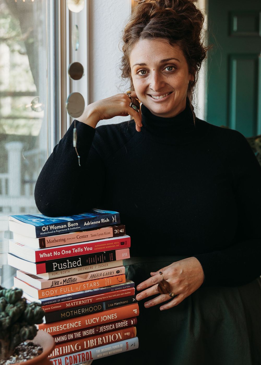 Someone wearing a black turtleneck sweater holds a stack of books while posing against a door in natural lighting.