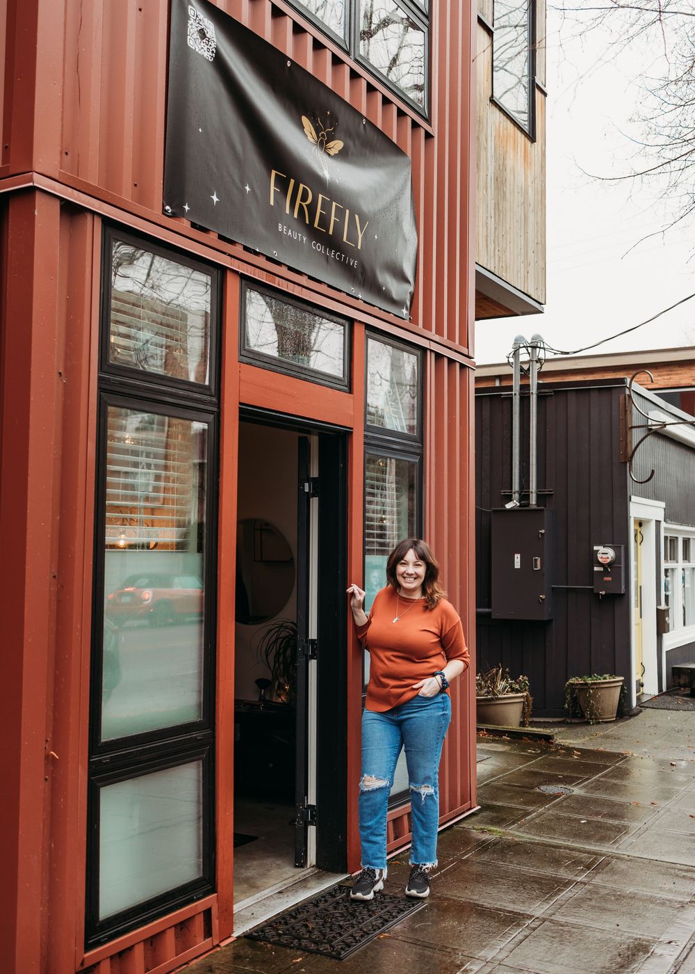 Modern retail storefront with container architecture featuring red metal siding and glass windows on a rainy day.