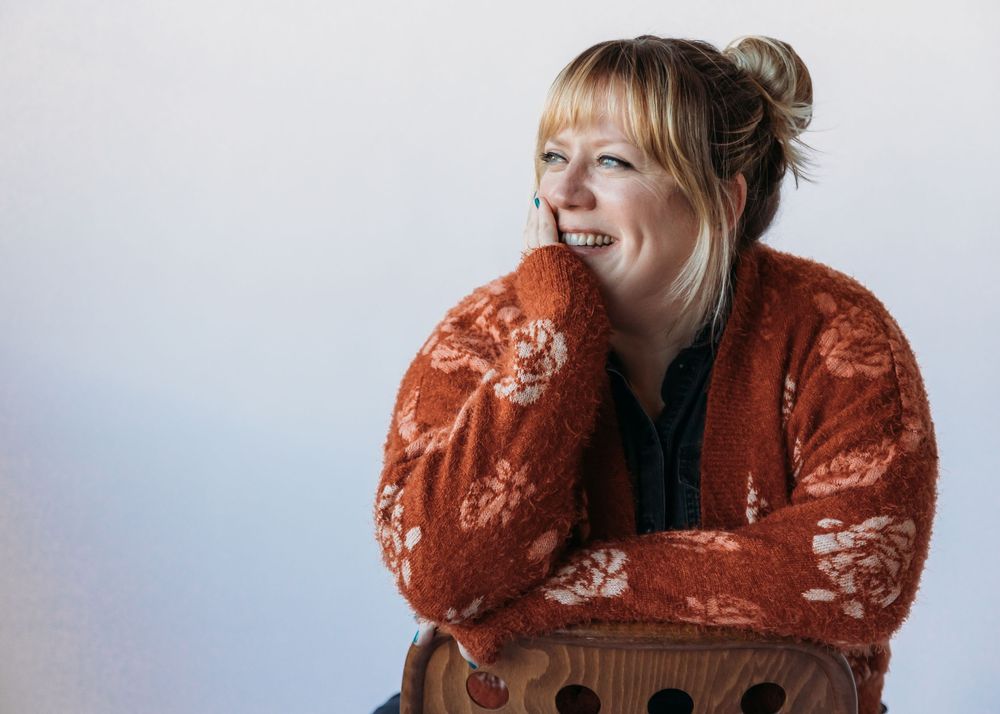 Person in burnt orange floral sweater sitting on wooden chair against light background, laughing with eyes closed.