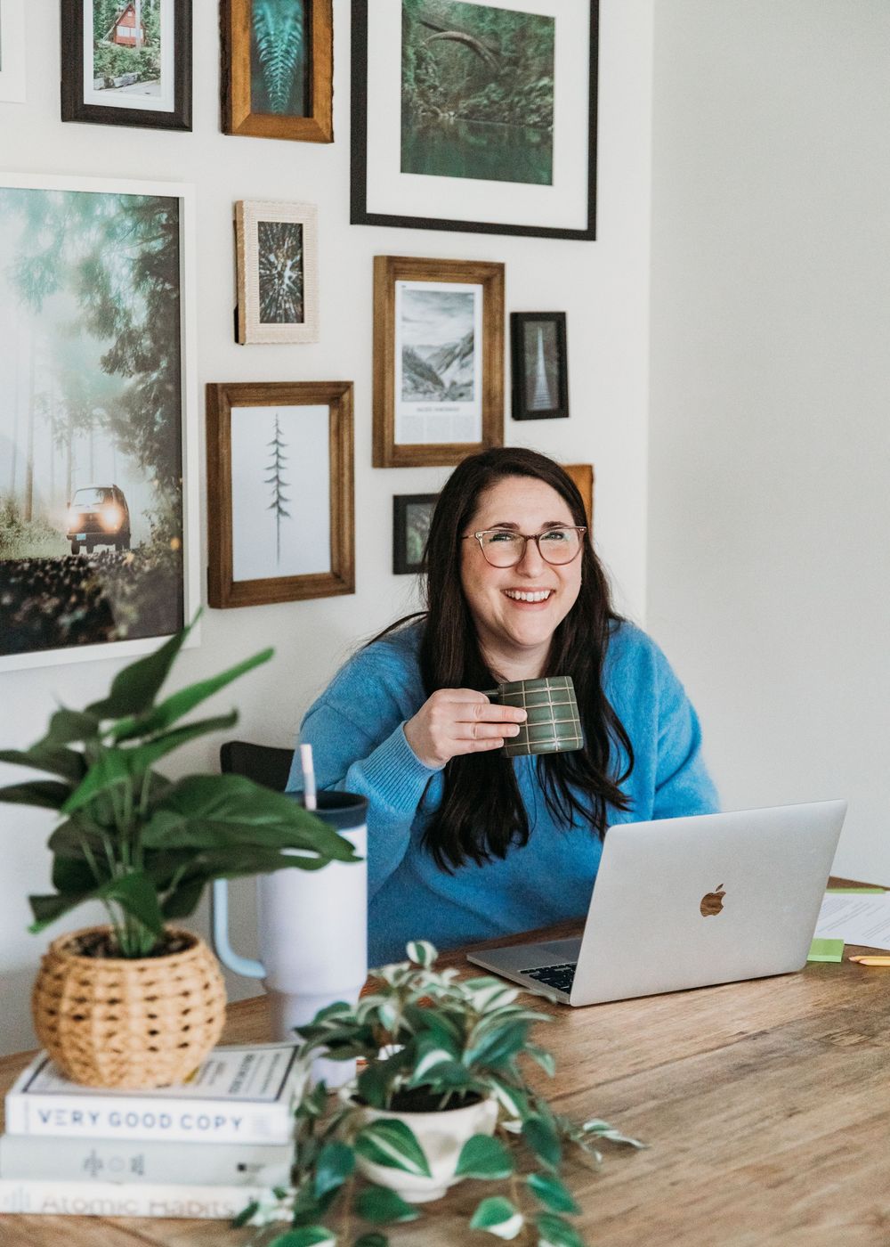 Cheerful remote worker in blue sweater sits at wooden desk with laptop surrounded by framed artwork and plants.