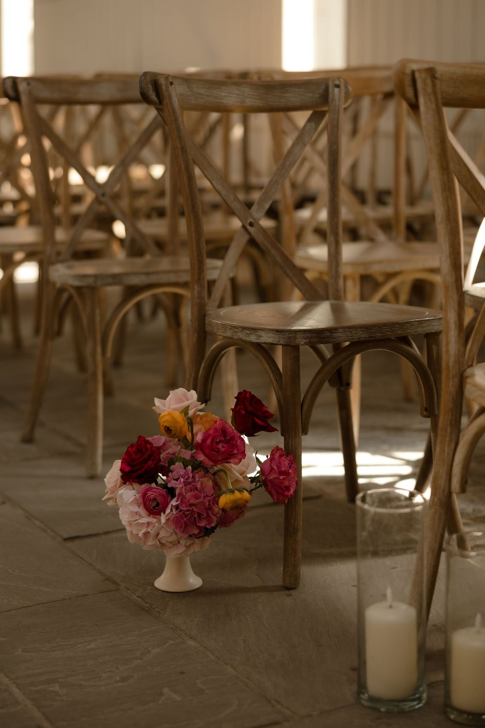 Rustic wooden cross-back chairs arranged with pink and burgundy floral centerpiece and white candles on stone floor.