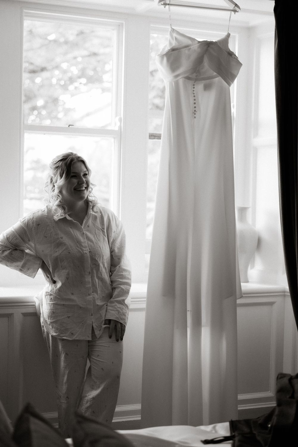A classic white wedding dress with button details hangs by a bright window in a simple, elegant monochrome setting.