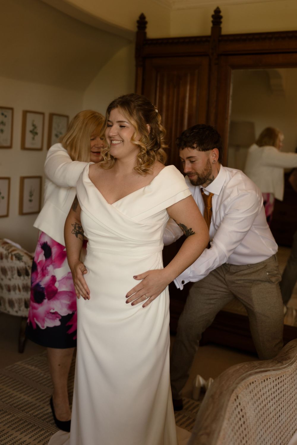 A wedding preparation scene shows a bride in an elegant white dress being helped by two attendants in a warm indoor setting.
