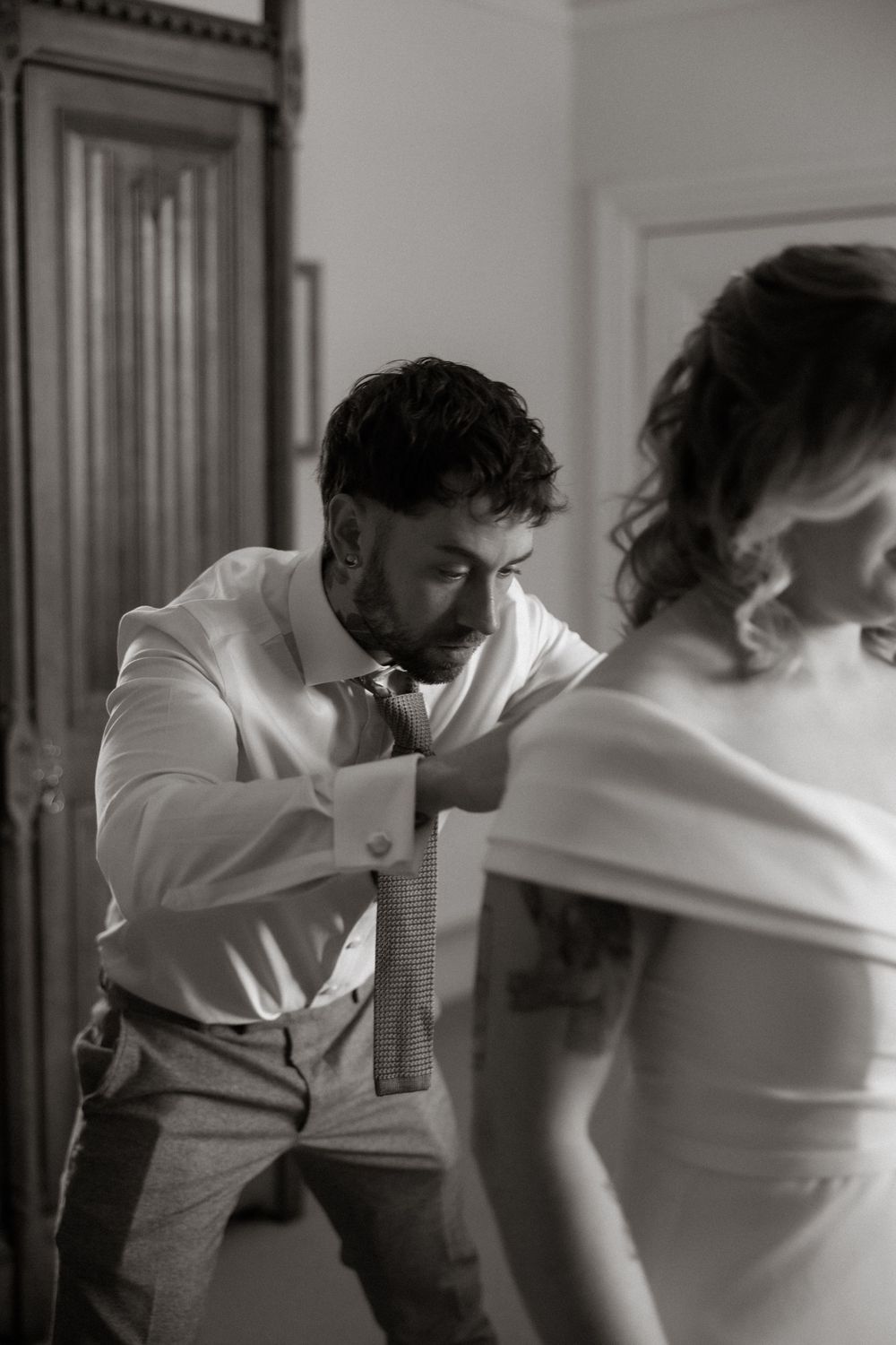 A groom helping with wedding preparations in a monochromatic scene wearing a white dress shirt and gray pants.