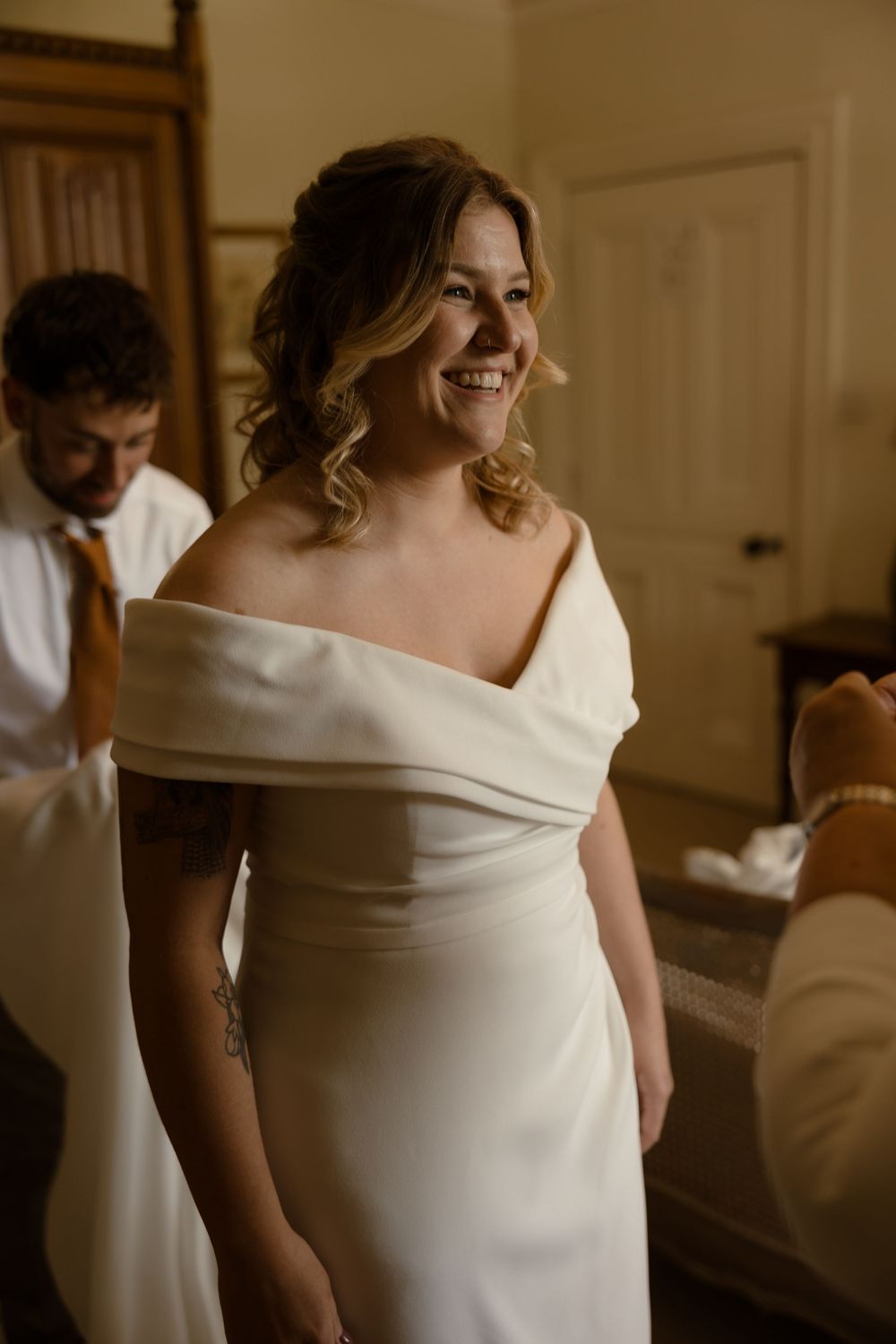 A bride in an elegant off-shoulder white wedding gown smiles warmly while getting ready in a sunlit room.