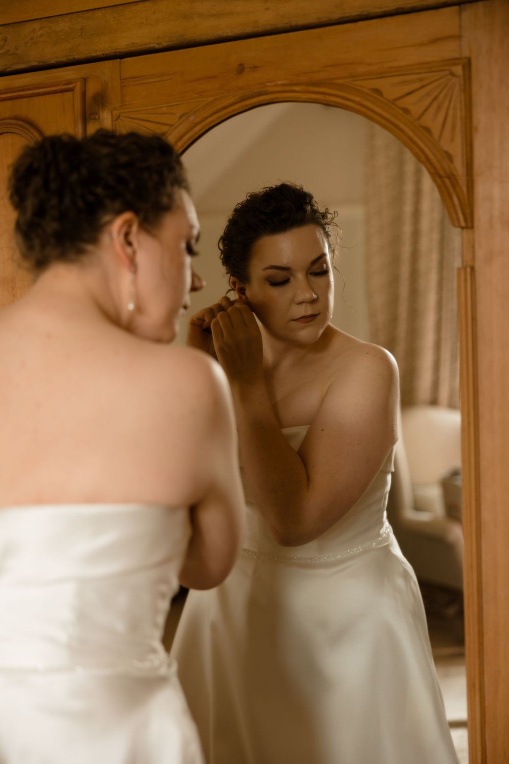 A person wearing a white wedding dress gazes thoughtfully into a wooden framed mirror while getting ready.
