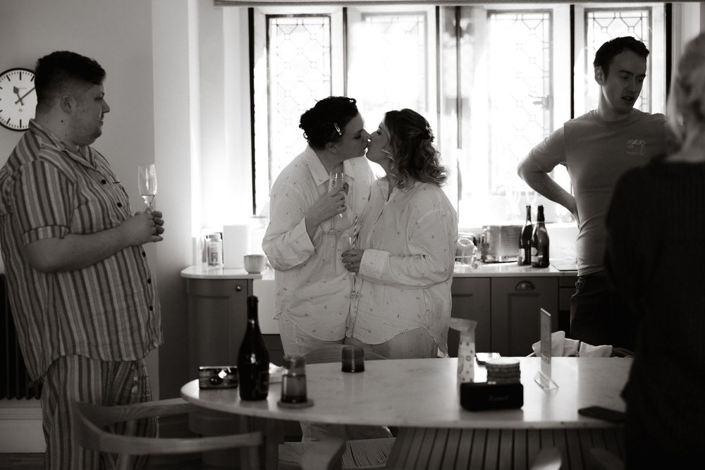 A black and white photograph shows people gathering in a casual indoor setting with drinks on a counter.