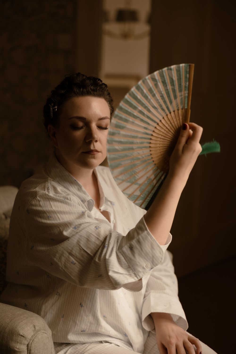 A figure in a vintage white dress holds an ornate paper fan in a moody, sepia-toned portrait.