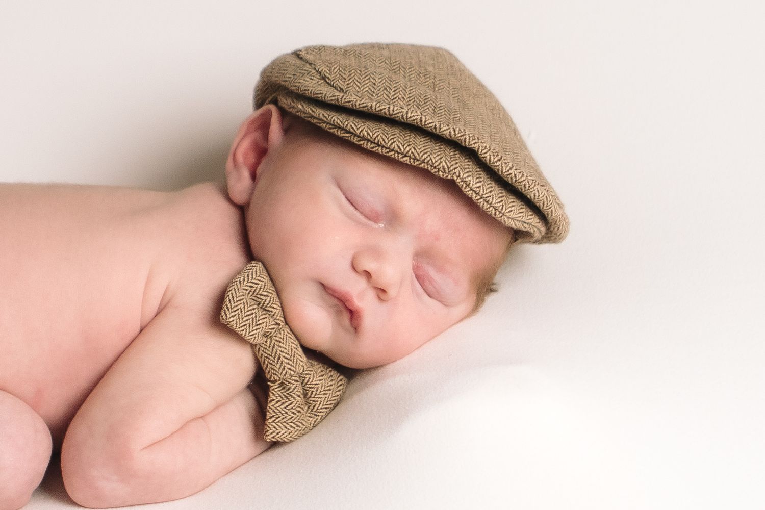 Sleeping newborn wearing tan newsboy cap and matching bow tie during an in home newborn session in Buffalo New York