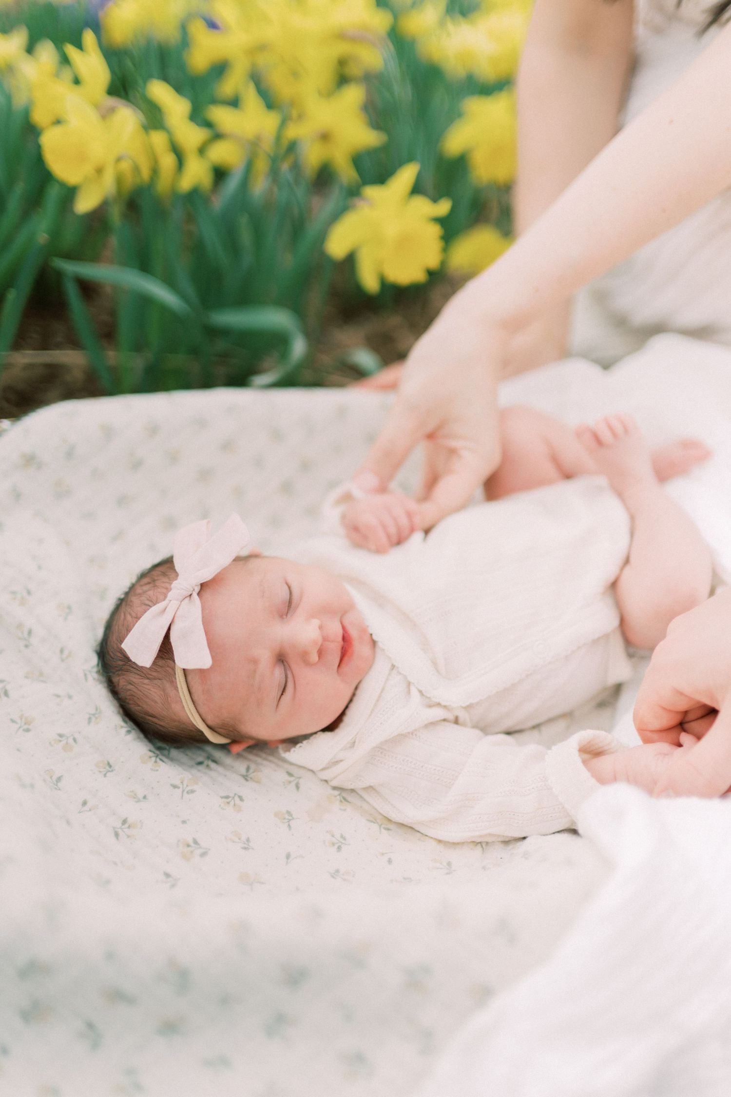 A baby in white clothing lies on a delicate blanket outdoors with daffodils blooming in the background.