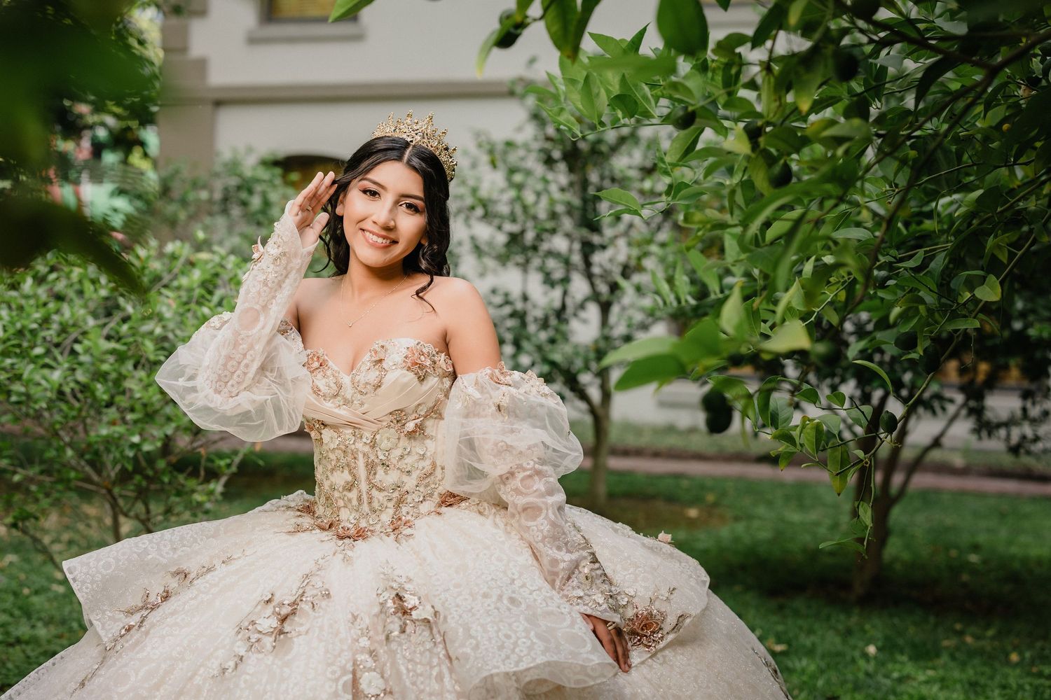Elegant quinceanera in a luxurious gold and white ball gown poses in a garden setting during golden hour.