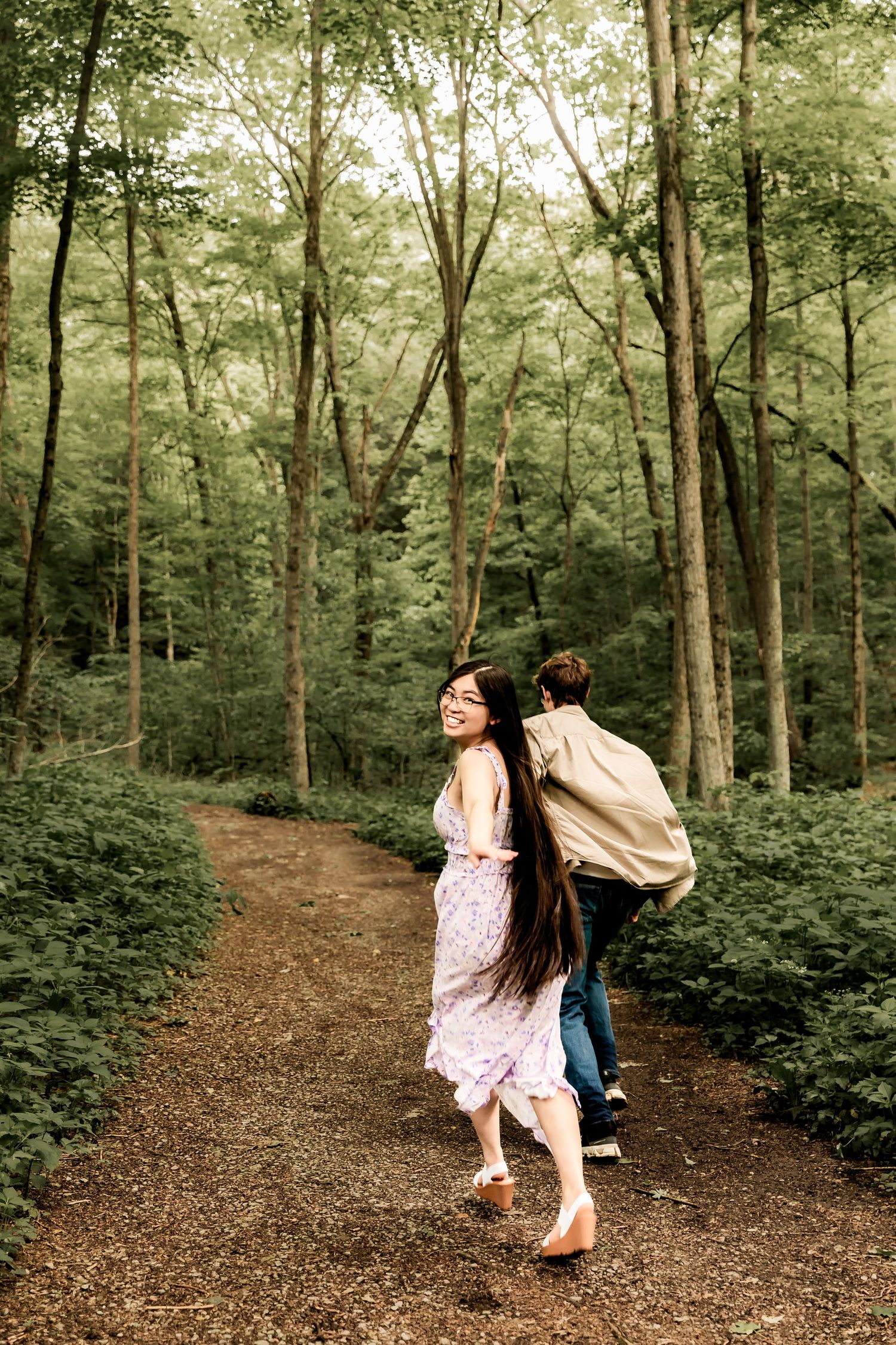 A couple walks along a scenic forest trail surrounded by tall green trees on a summer day.