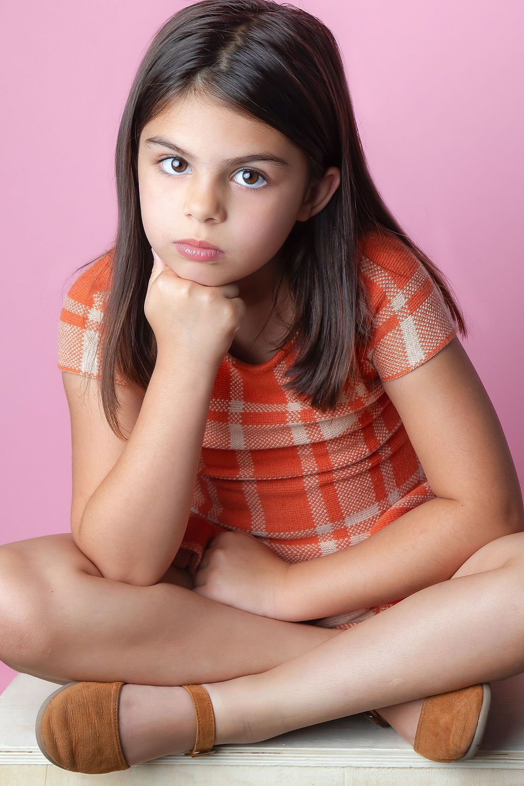 Young person in orange plaid shirt sitting cross-legged with a contemplative expression against pink background.