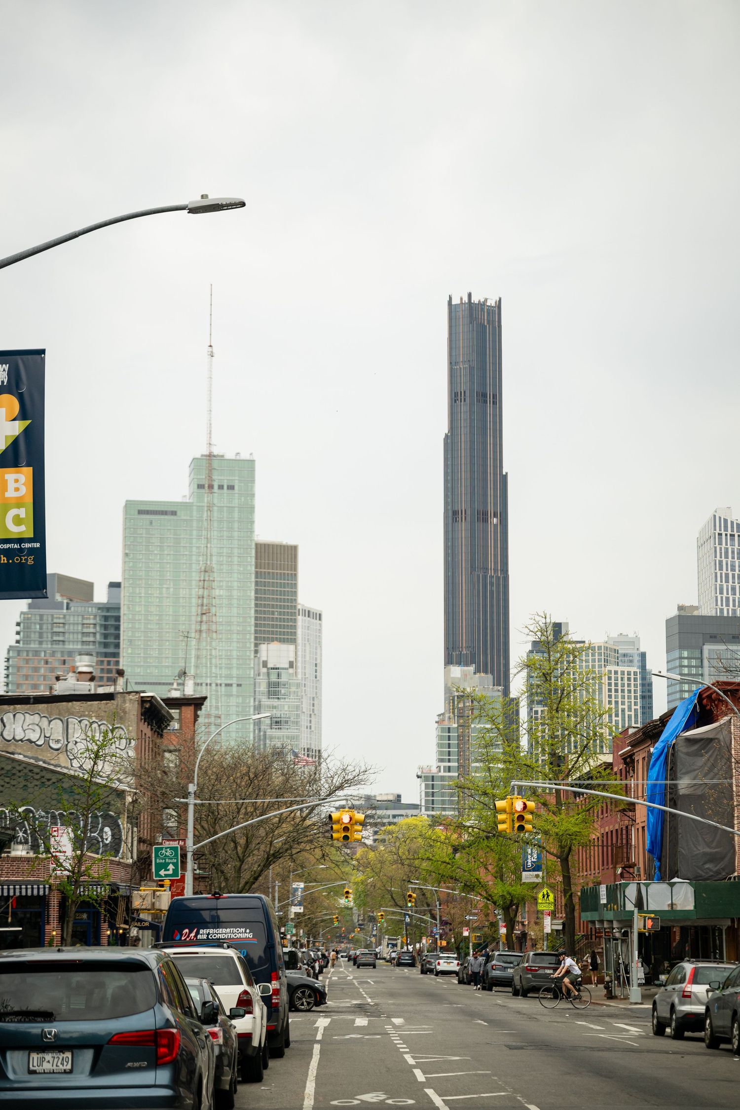 Urban street view in New York City with tall skyscrapers including a prominent dark tower against an overcast sky.