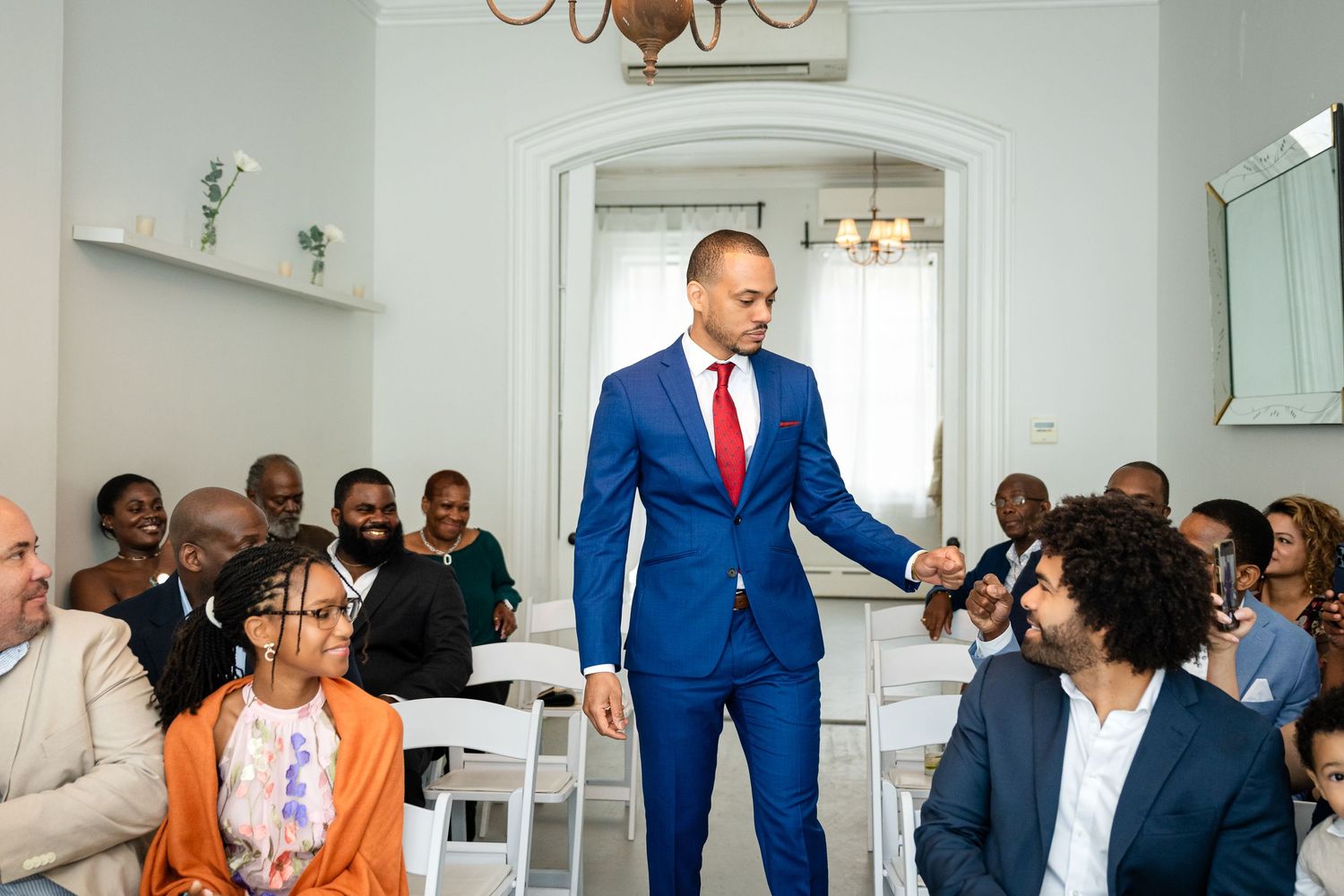 People gather in a bright white room as a man in a vibrant blue suit stands and gestures during an event.