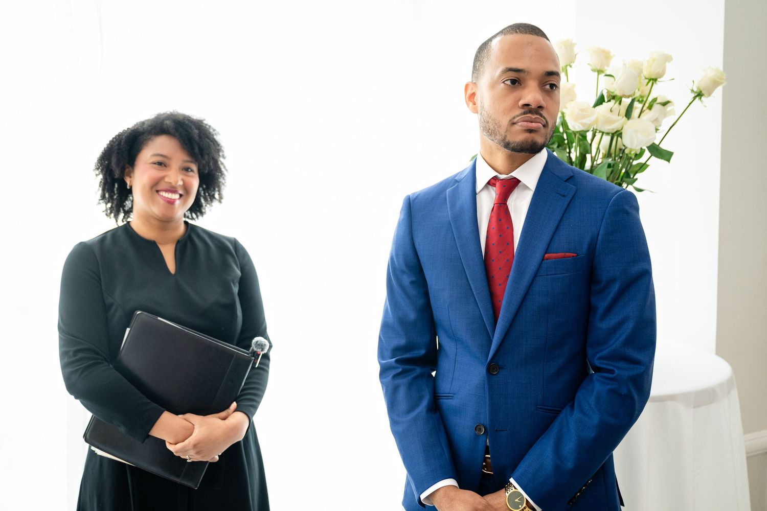 Two business professionals in professional attire stand together in a bright office setting with white flowers in background.