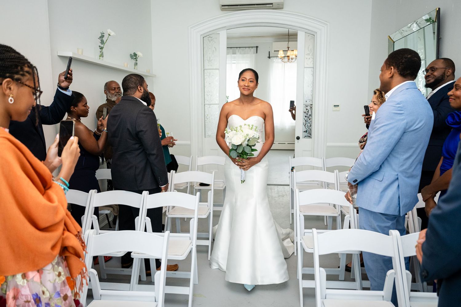 A bride in a white strapless wedding gown stands in a bright white room filled with white folding chairs and wedding guests.