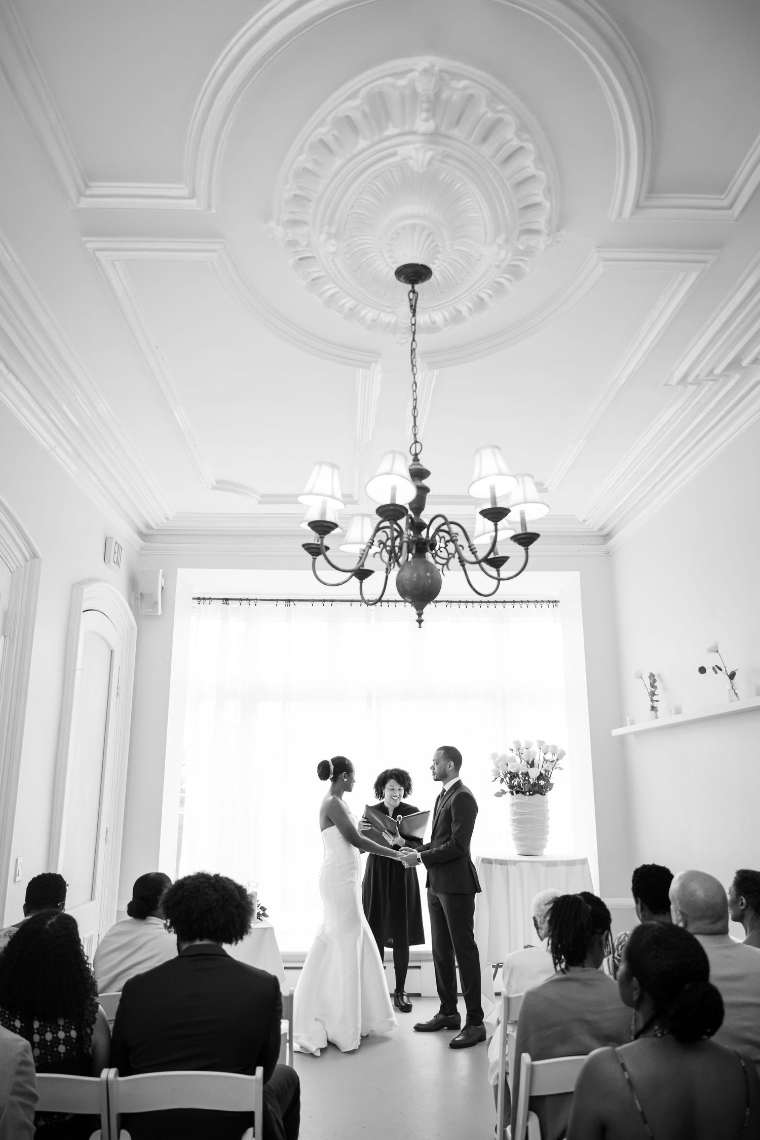 A wedding ceremony takes place in a bright white room with an elegant chandelier and ornate ceiling medallion.