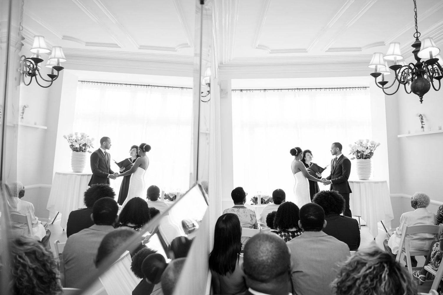 Guests watch a wedding ceremony in a bright white circular room with elegant chandeliers and vaulted ceilings.