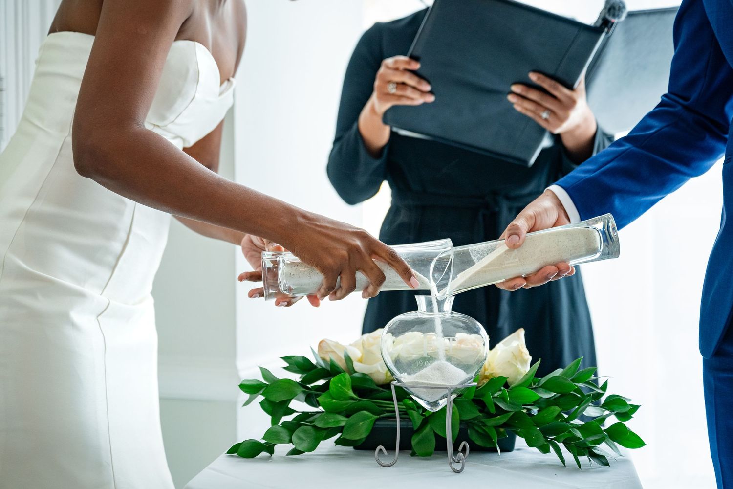 A couple performs a unity sand ceremony with white and green floral decor while officiant holds ceremony book.