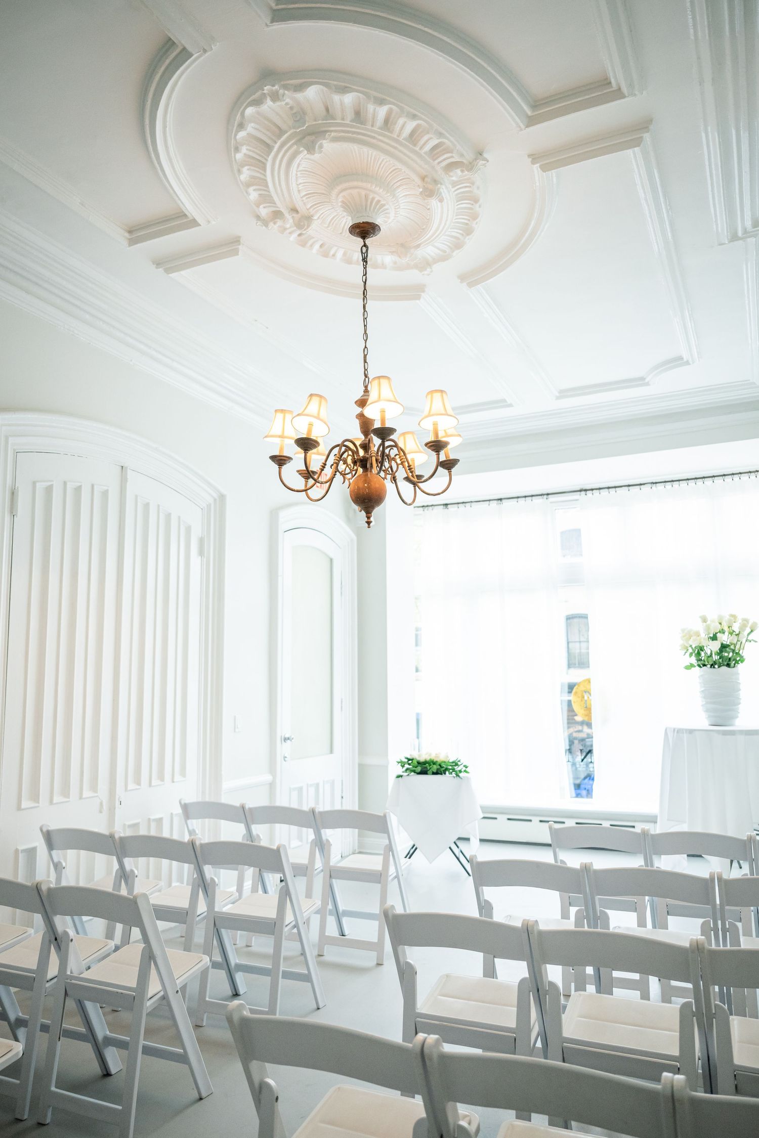 Interior of an elegant white wedding venue with folding chairs and an ornate chandelier under decorative ceiling molding.