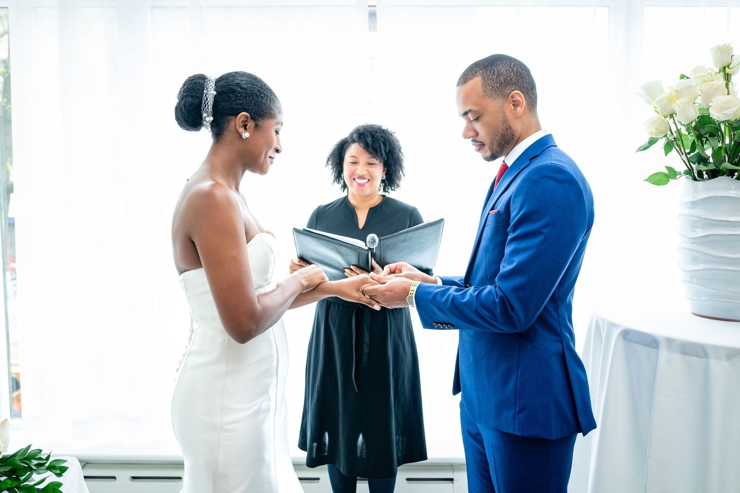 A couple stands at a wedding ceremony with an officiant in black robes as they exchange rings during their vows.