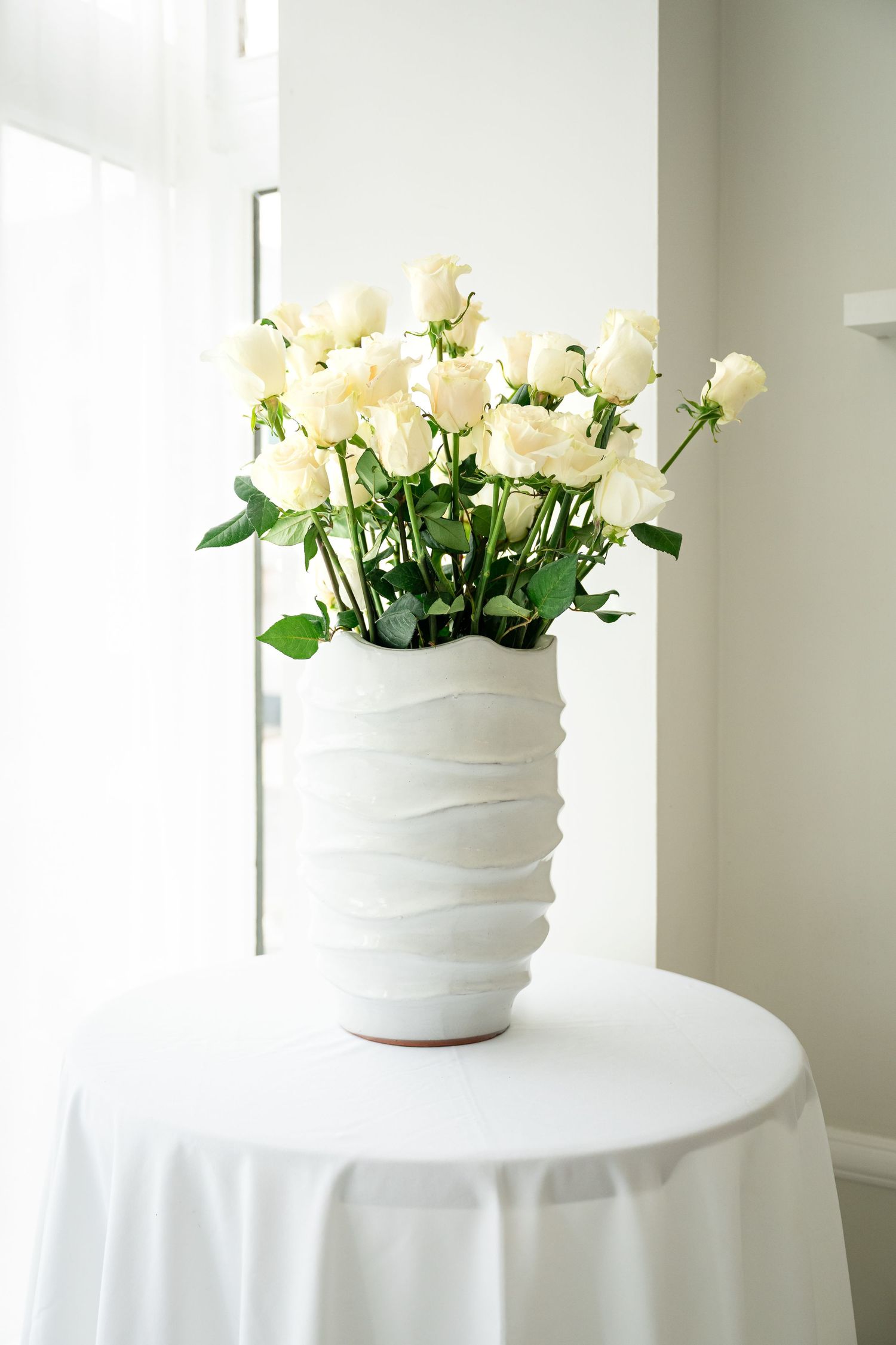 A bouquet of creamy white roses arranged in a modern textured white ceramic vase sits atop a round table with white tablecloth.