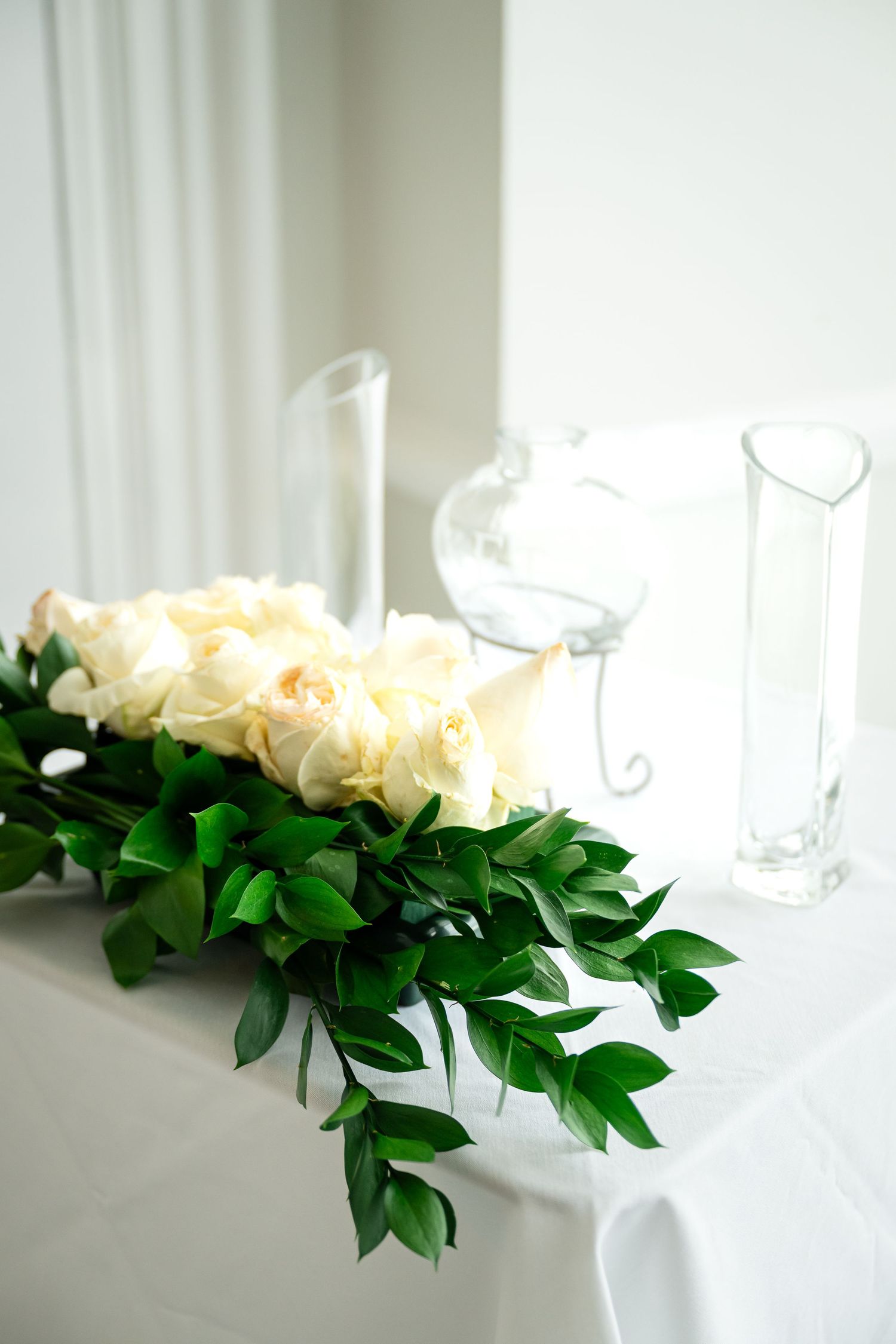 White roses and green leaves arranged on white table with clear glass vases creating an elegant tablescape setting.