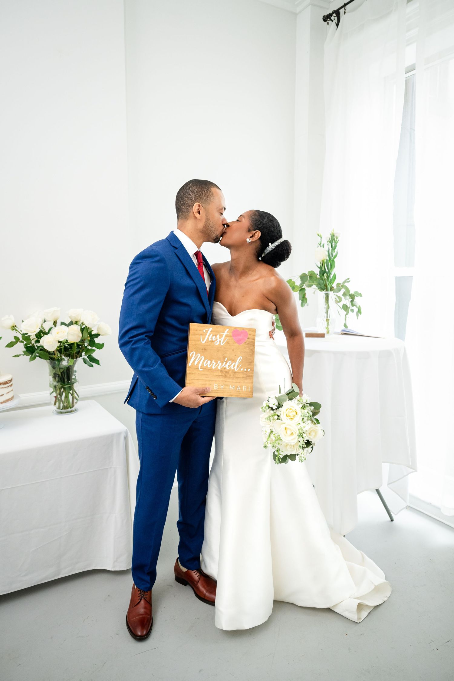 A wedding couple sharing a romantic kiss in a bright white room with floral decorations.