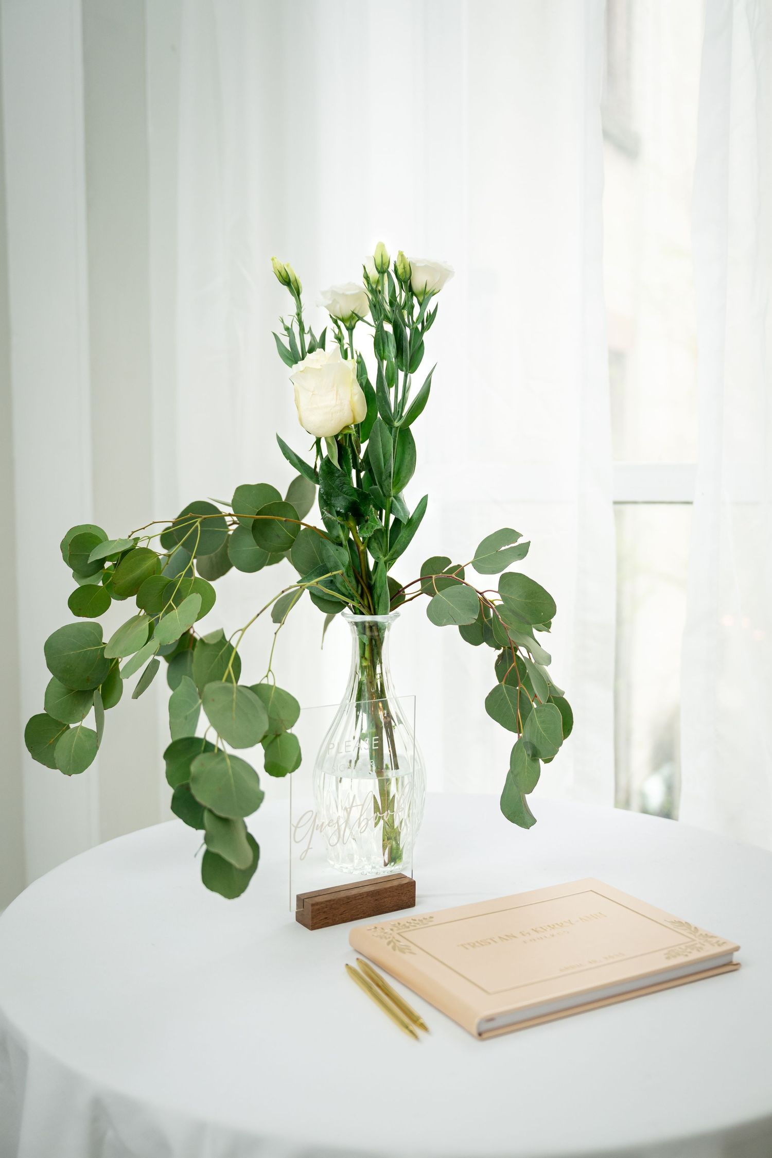 A minimalist vase with white flowers and eucalyptus leaves sits on a white table next to a notebook and pencil.