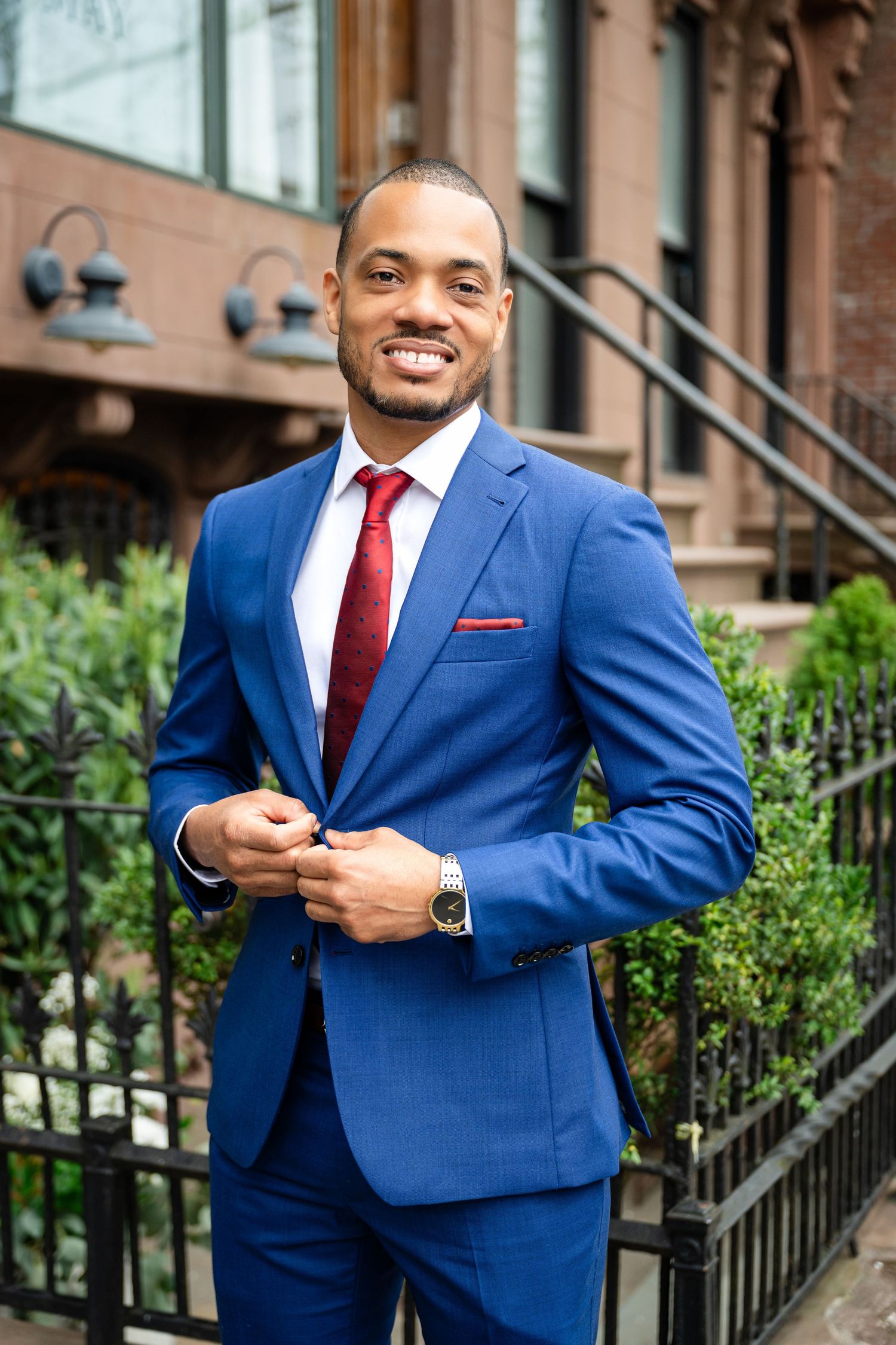 A professional in a tailored bright blue suit with red tie stands smiling outside a brownstone building with wrought iron railings.
