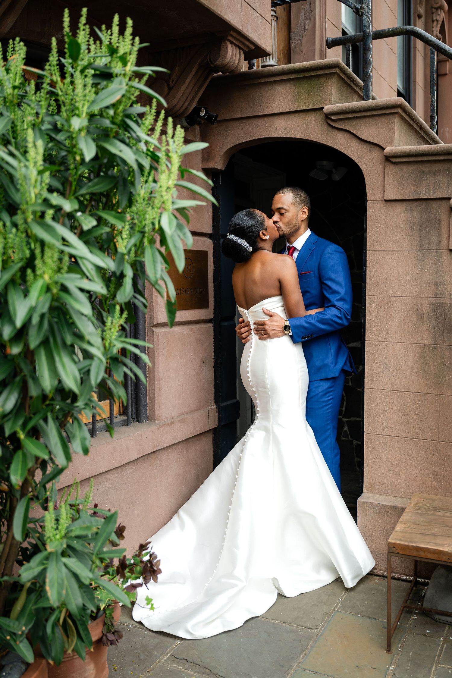 Newlywed couple shares romantic moment in formal attire by brownstone entrance.