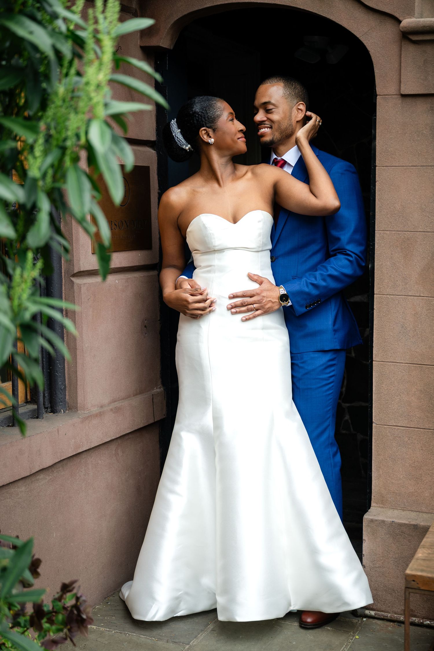 Couple in wedding attire embraces in archway of brownstone building.