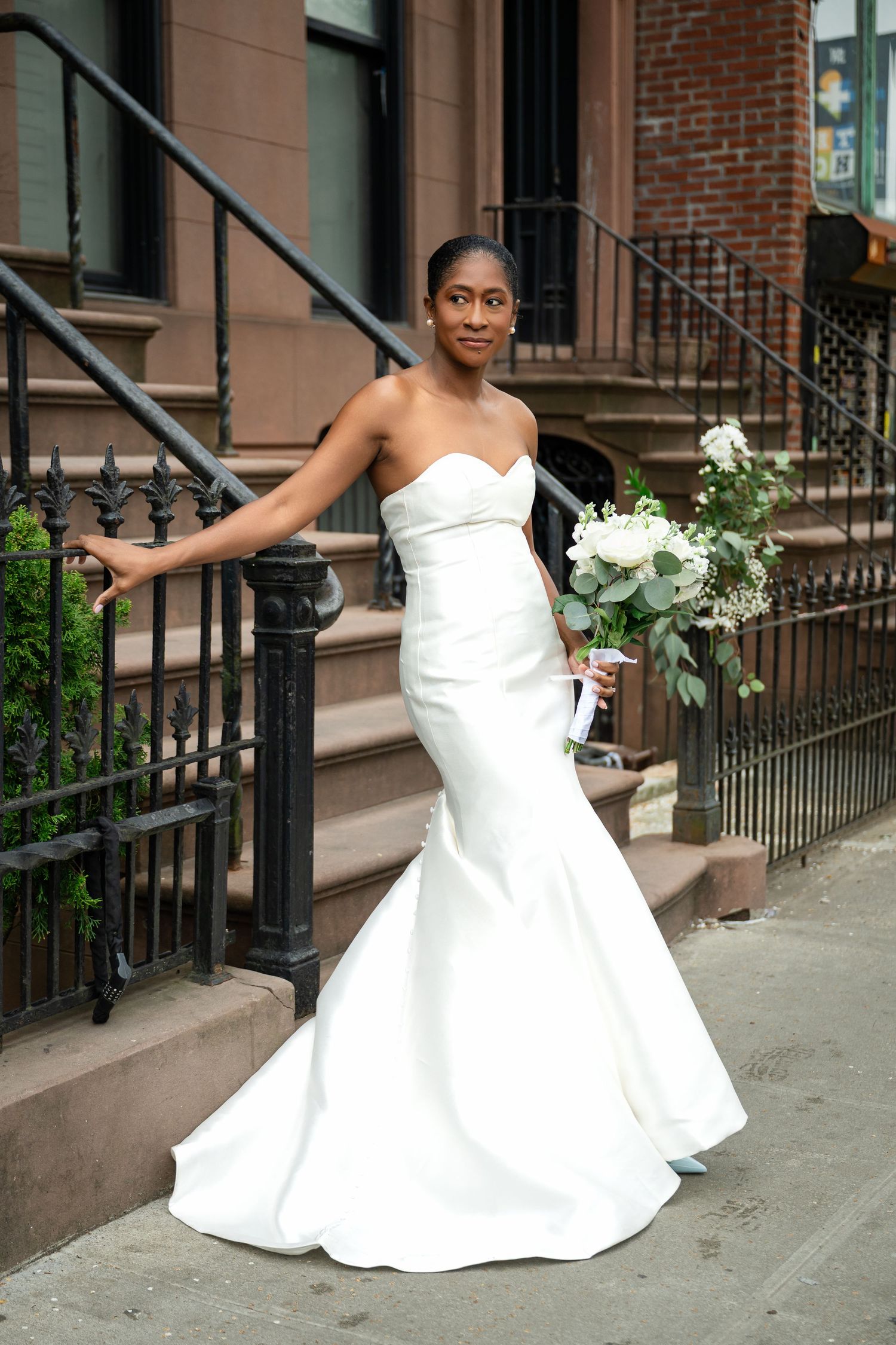 A woman in an elegant white strapless mermaid wedding gown holds a bouquet while standing on brownstone steps.