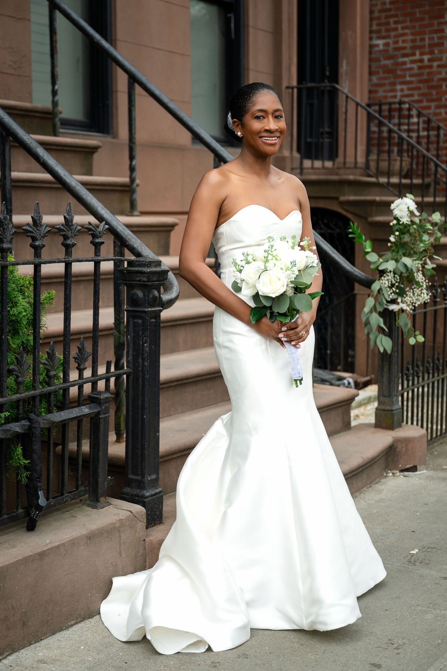 Bride in elegant white mermaid wedding dress holding a bouquet on brownstone steps.