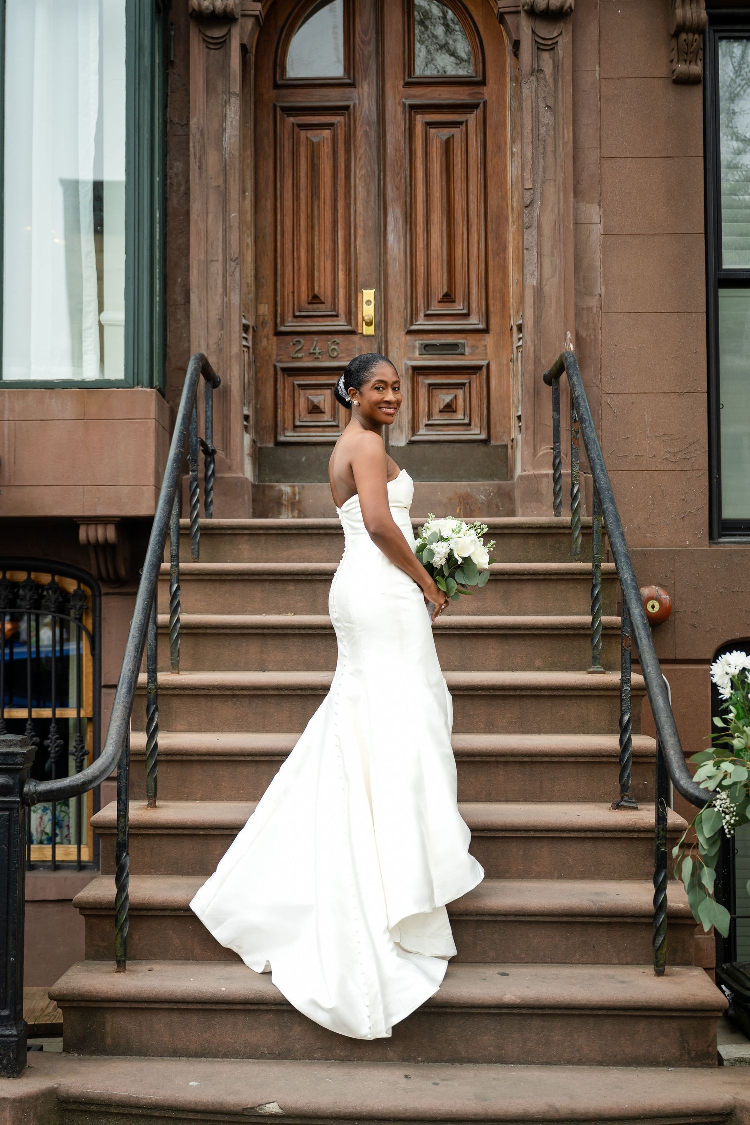 A bride in a fitted white gown poses on brownstone steps in front of an ornate wooden door.