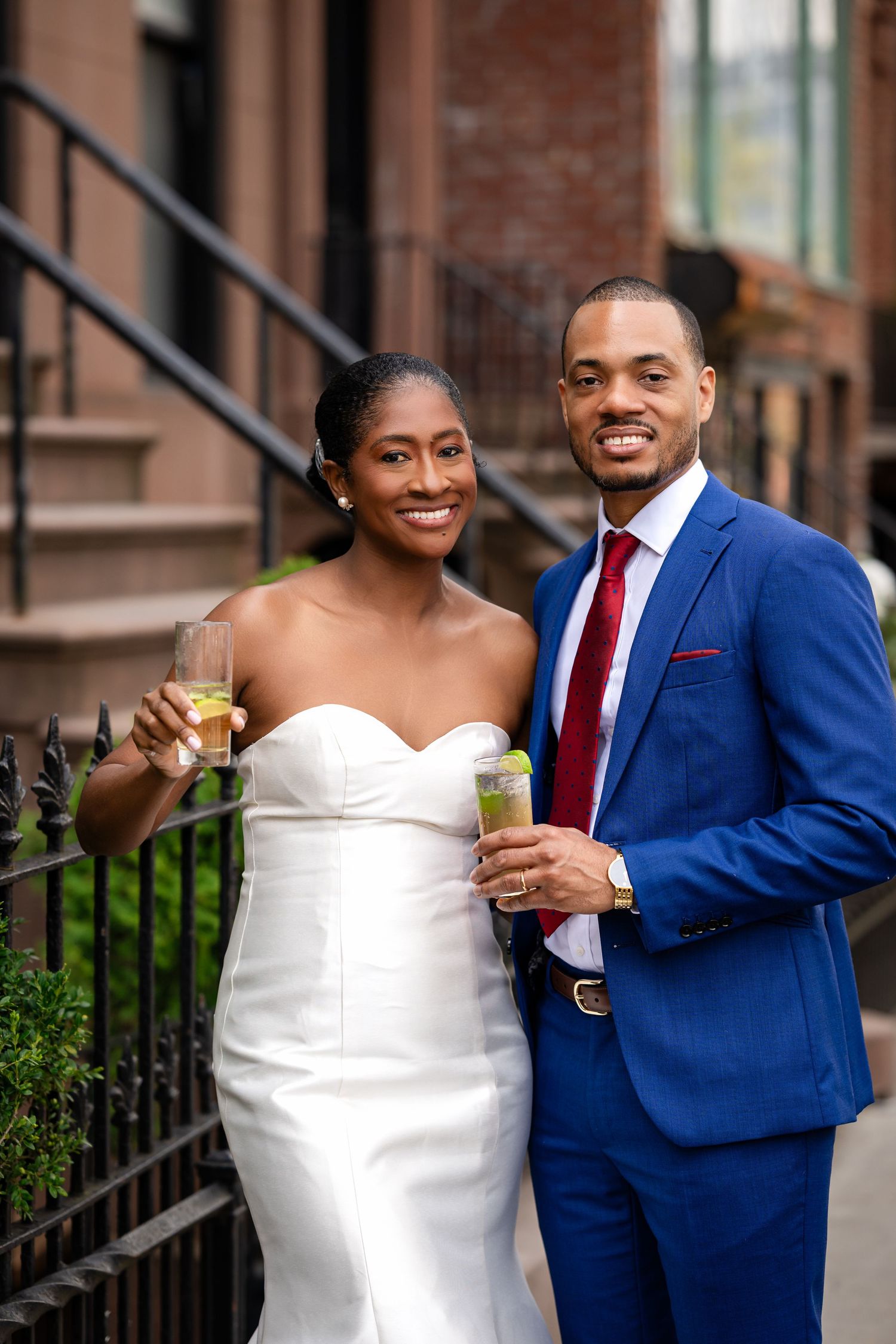A couple in wedding attire holding drinks and smiling on brownstone steps.