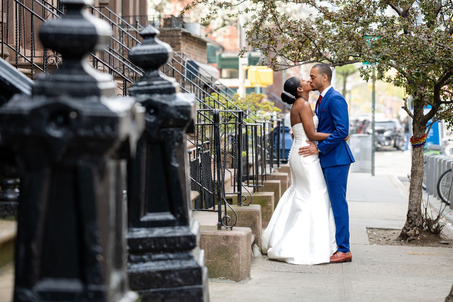 A wedding couple embraces on a city sidewalk with classic brownstone architecture in the background.