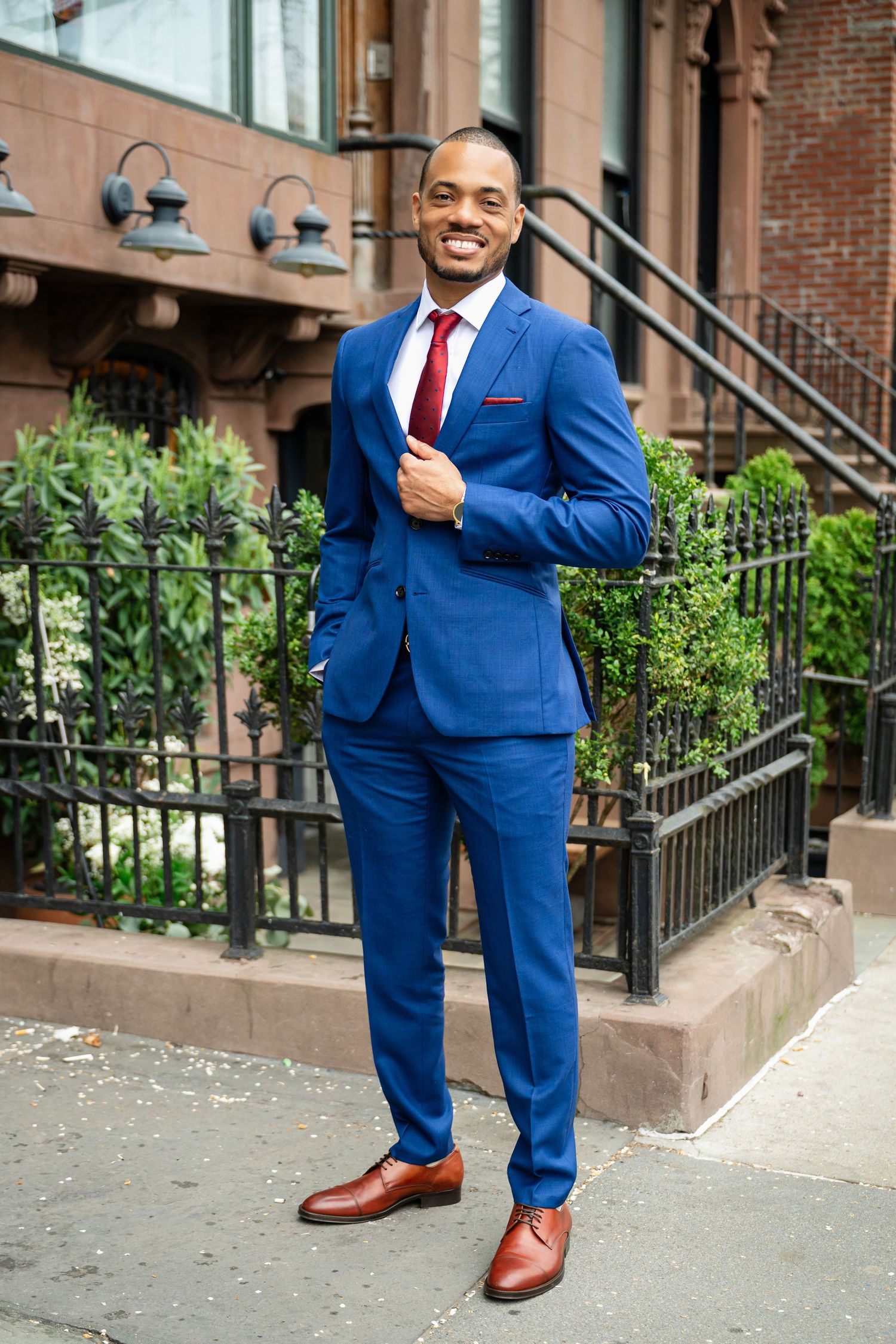 A person in a bright blue suit and red tie stands confidently on a city sidewalk near brownstone steps and iron railings.