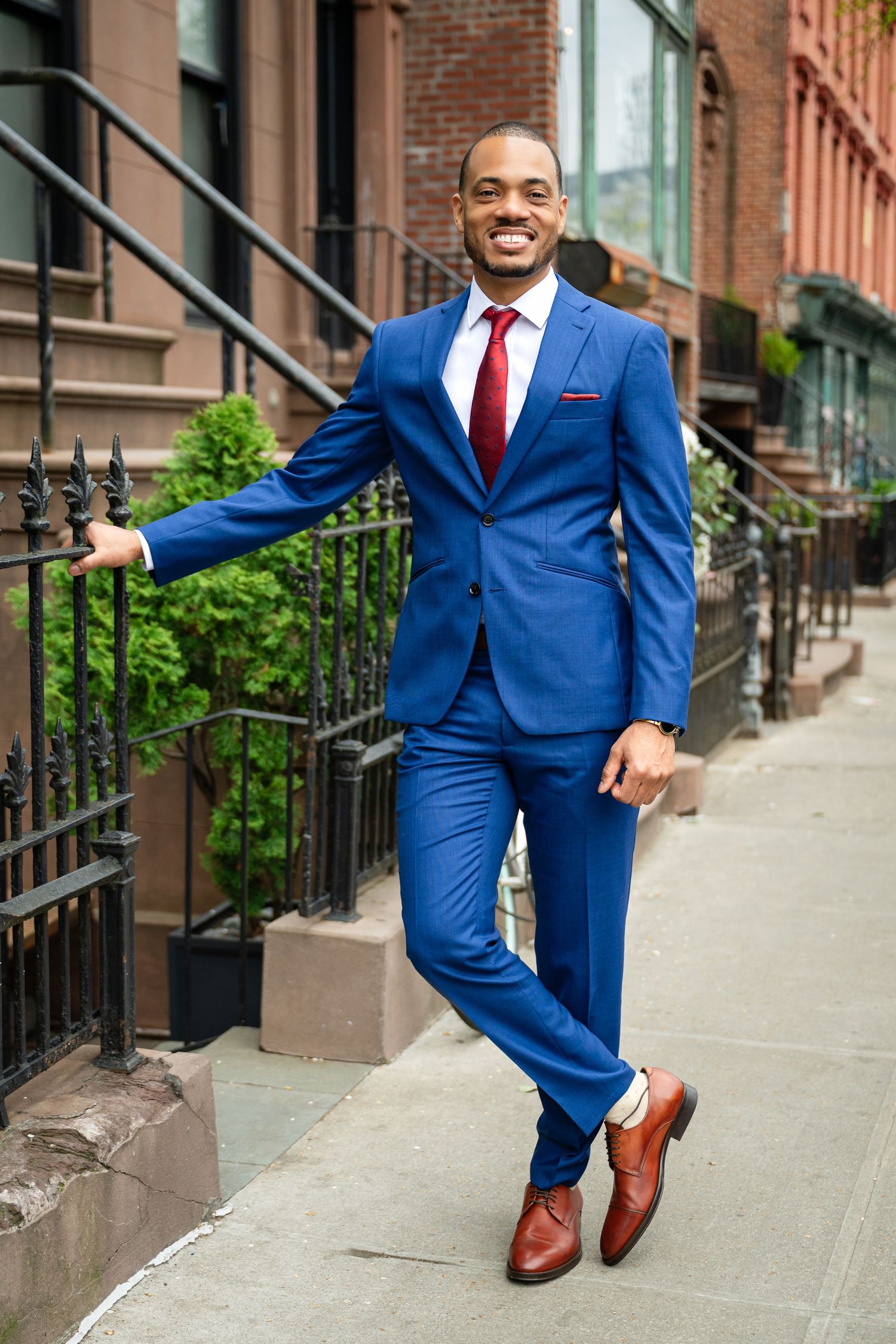 A person in a tailored blue suit and red tie poses confidently on a city brownstone staircase.