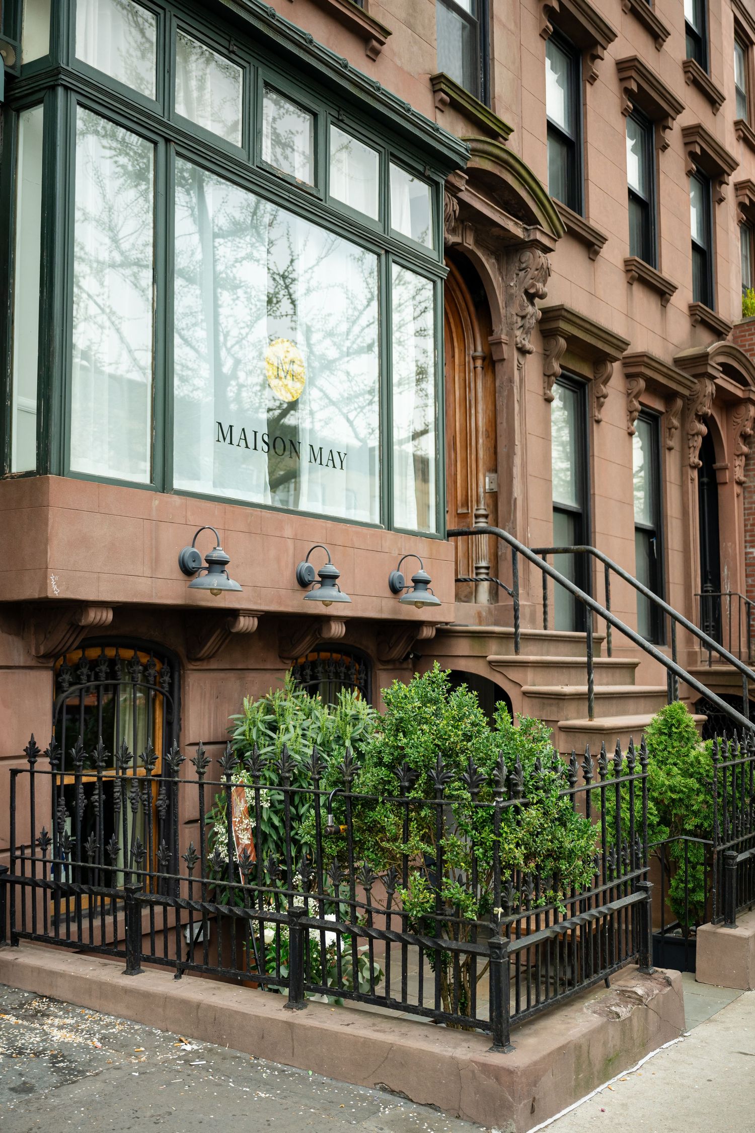 Brownstone storefront entrance with green-framed windows and 'Maison May' signage along a Brooklyn street with wrought iron railing.