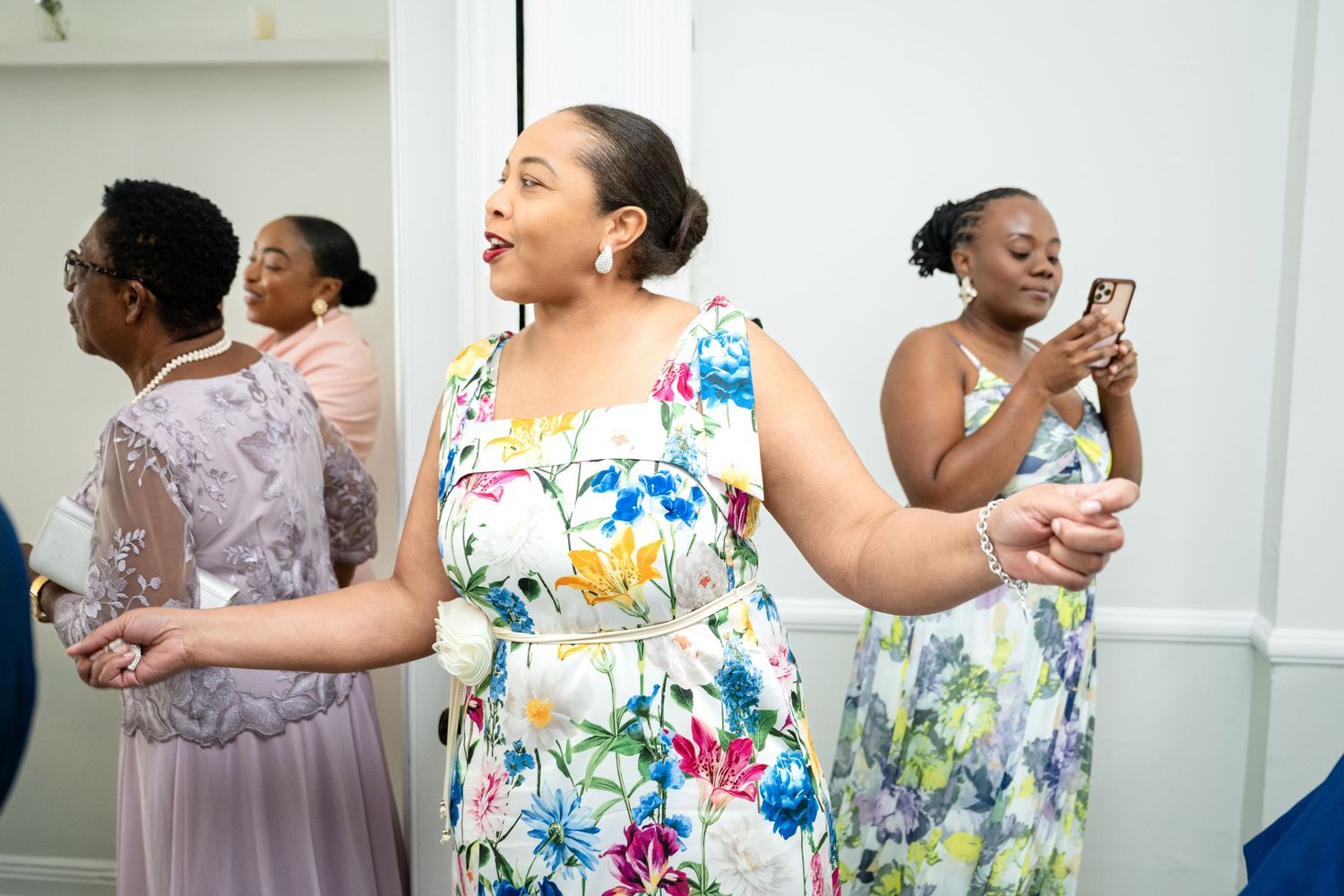 Guests in colorful floral dresses smile and laugh while standing together at a social gathering.
