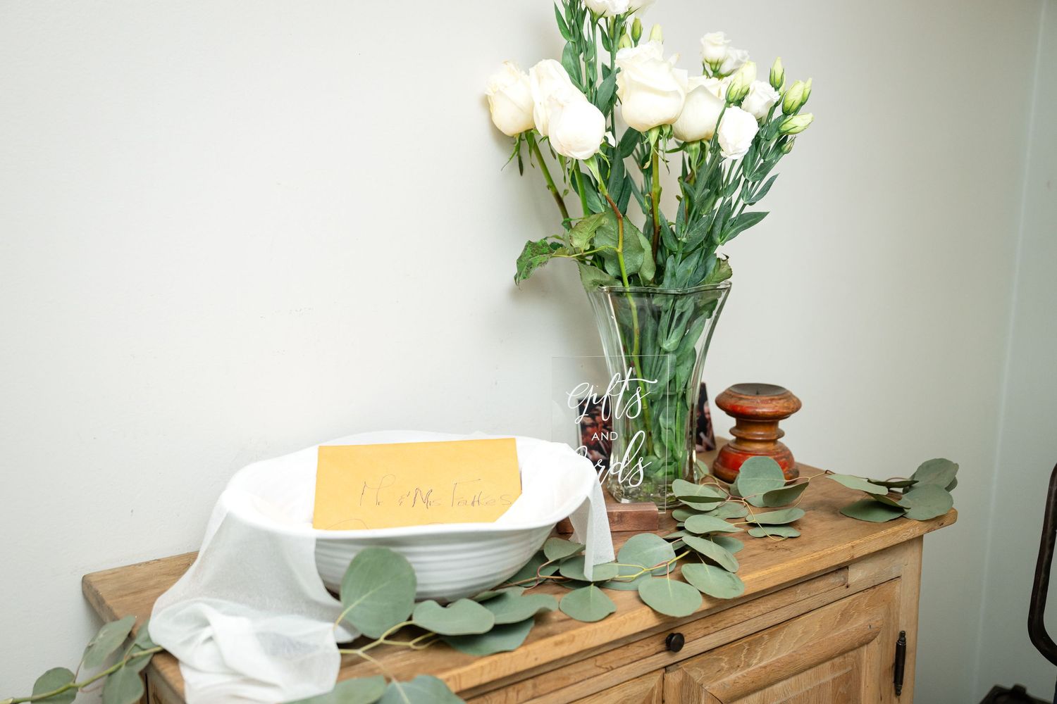 White roses in a glass vase with eucalyptus leaves on a wooden surface.