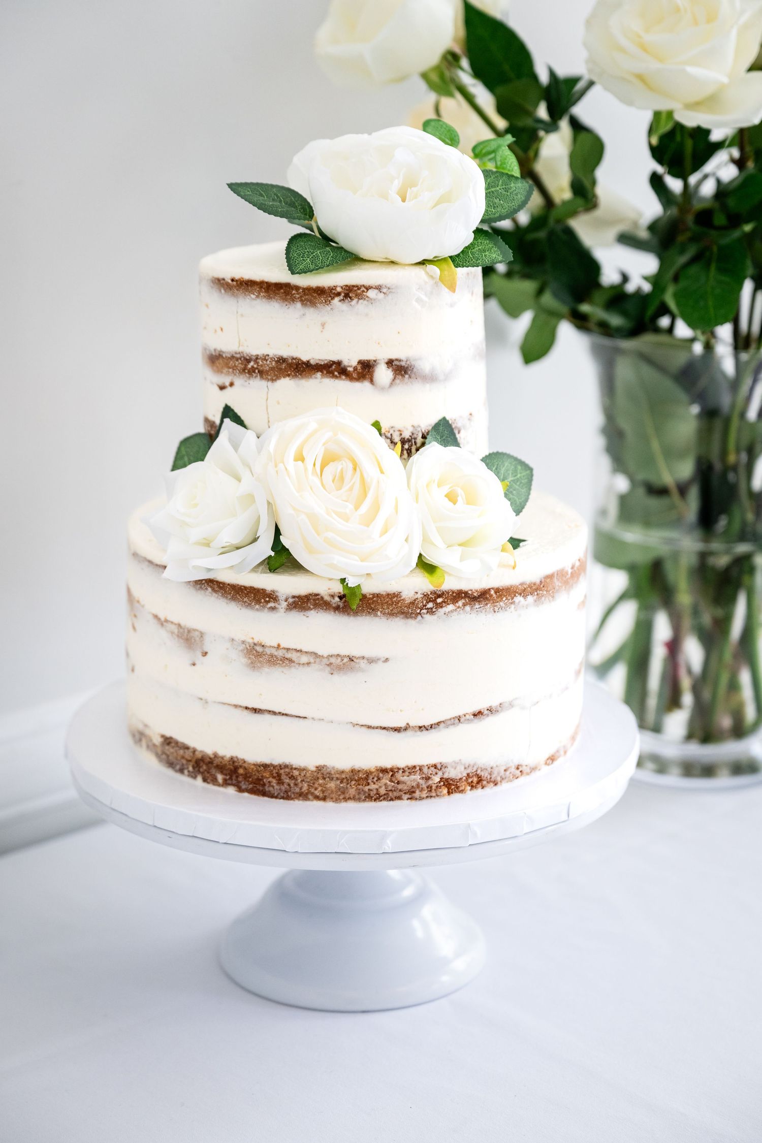A multi-tiered naked wedding cake decorated with white roses on a white cake stand against a light background.