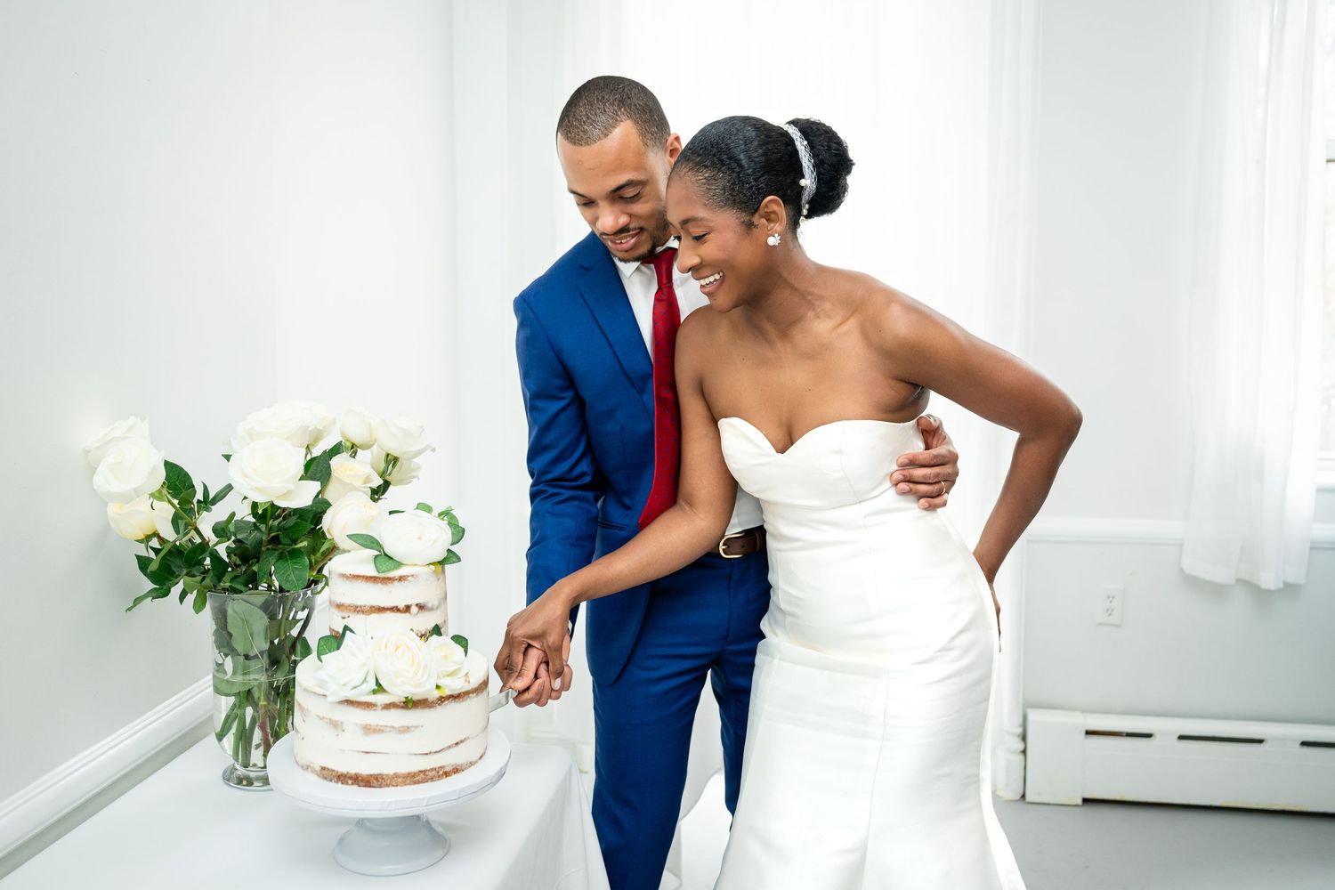 A couple cuts their elegant white wedding cake together beside a bouquet of white flowers.