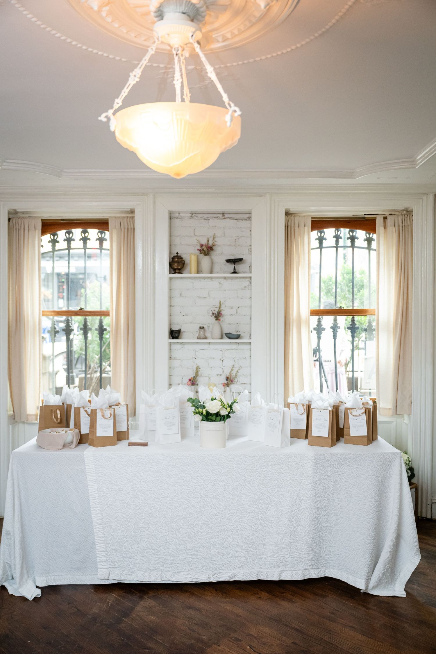 Elegant white dining table setup with chandelier and tall windows in bright room.