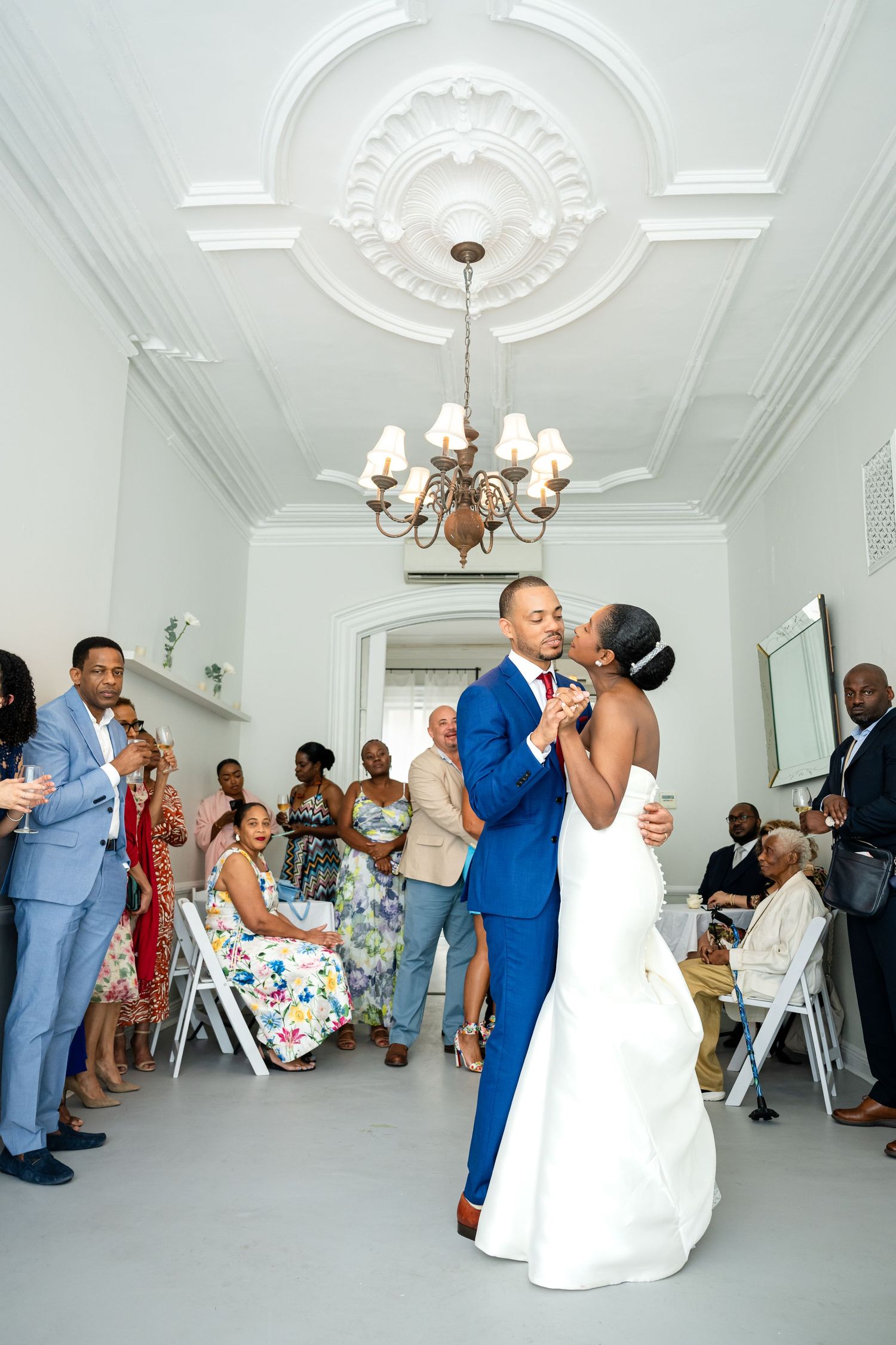 Wedding couple shares their first dance under an ornate chandelier in a bright white room.