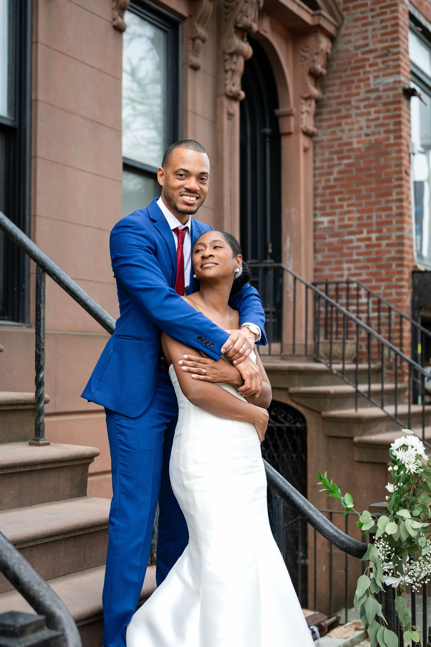 Newlyweds embrace on brownstone steps with brick building and white flowers in background.