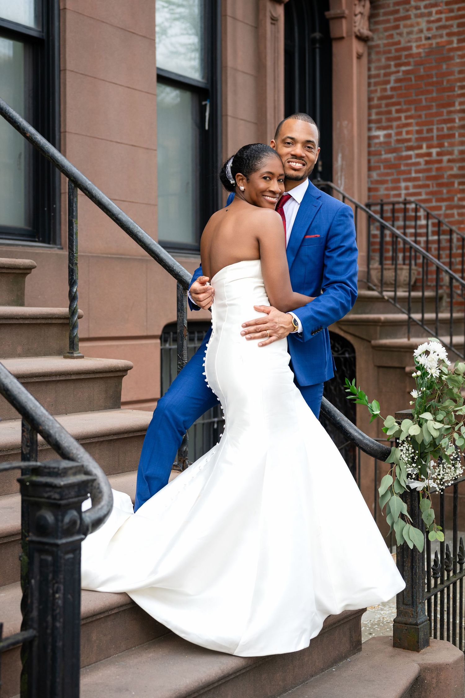 A couple in their wedding attire pose on brownstone steps in front of a brick building.