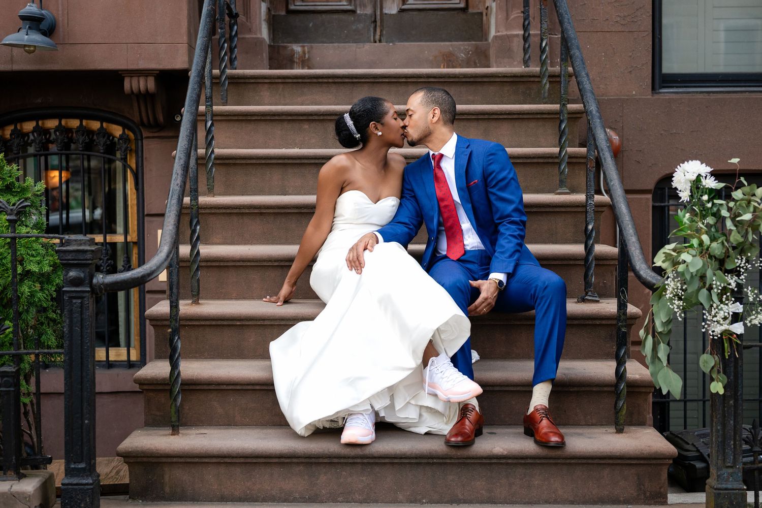 A joyful wedding couple sharing a moment while seated on brownstone steps decorated with floral arrangements.